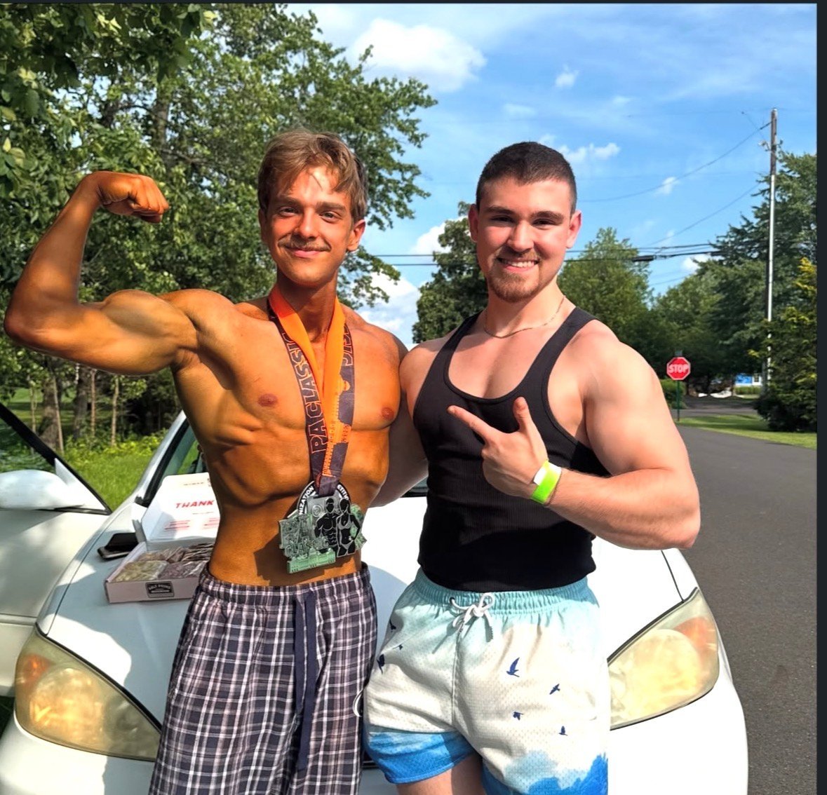 Lance posing outdoors by a white car with his bodybuilding client Ben, both smiling and flexing at the camera after a show